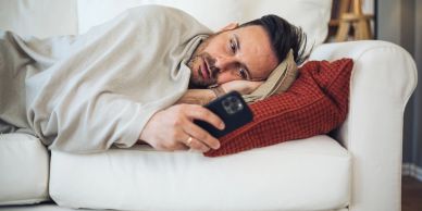 Man ligt op de bank met een deken en kijkt naar zijn telefoon.