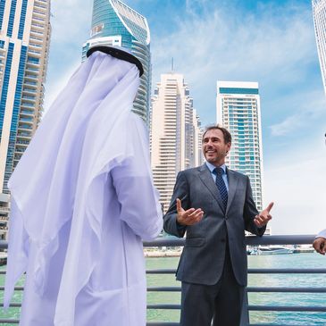 Three businessmen, two in traditional Middle Eastern attire, discussing outdoors near water and skyscrapers.