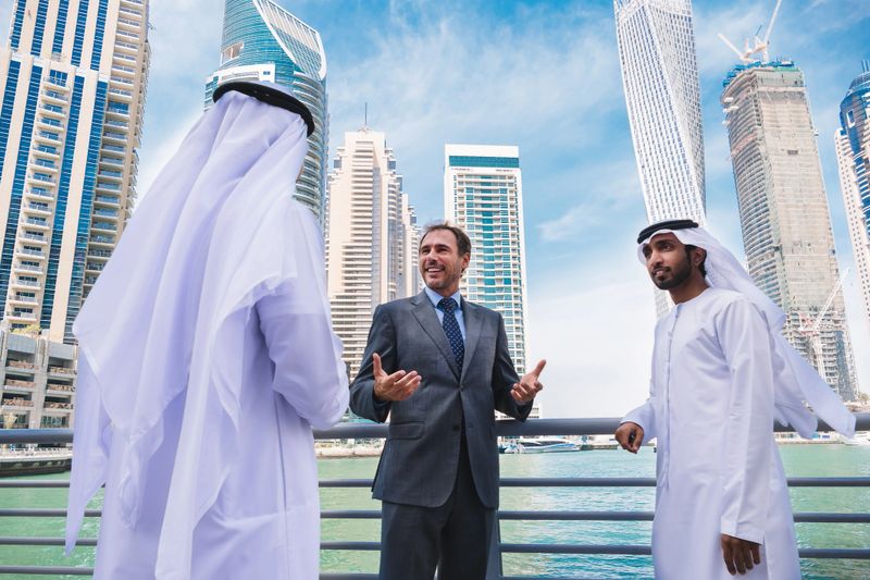 Three professionals, two in traditional attire and one in business suit, actively engage in a discussion near a river with striking skyscrapers in the background, showcasing the cohesion between modern business practices and cultural values set against a vibrant urban landscape.