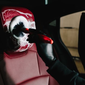 A person cleans red leather car seat with foam and brush.