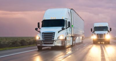 Two white semi trucks driving on a wet road at sunset.