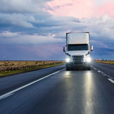 A large truck with headlights on driving on an open highway at dusk.