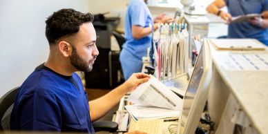 Man in scrubs working at a computer in a medical setting.