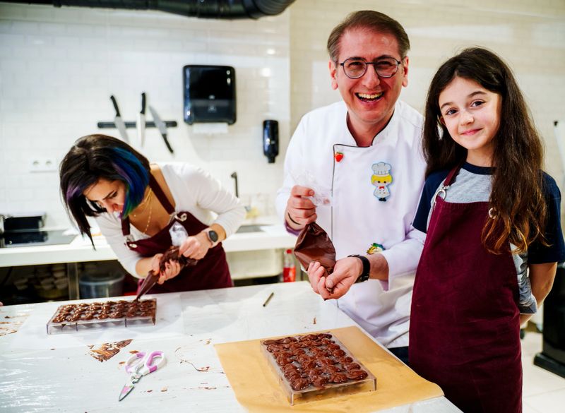 A heartwarming moment in a professional kitchen during a chocolate-making workshop. A male chef is guiding a child as they pipe melted chocolate into molds together. Both are wearing aprons, and the atmosphere is warm and focused. In the background, other participants are also engaged in the same activity. Perfect for concepts like culinary education, parent-child learning, food workshops, hands-on cooking classes, and teamwork in the kitchen.