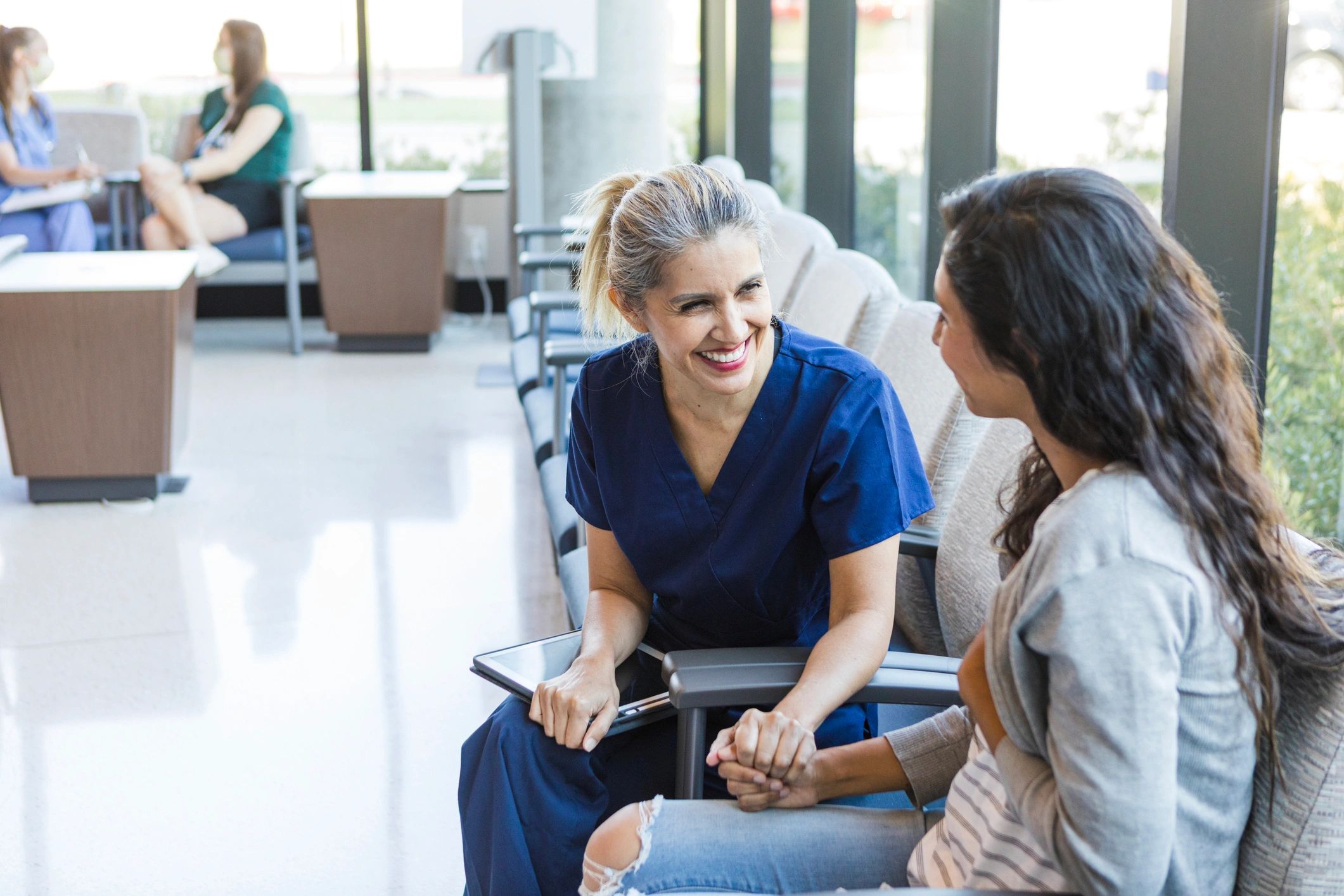 A nurse warmly holds a patient's hand in a bright waiting room.
