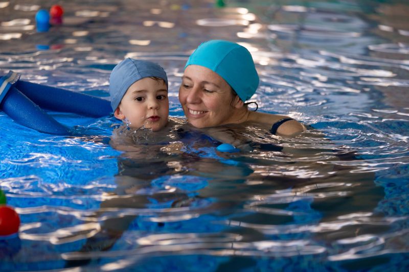 Mother teaching her son to swim, both wearing swim caps and smiling, in a pool with blue water