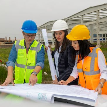 Three humans in hard hats discussing blueprints.  