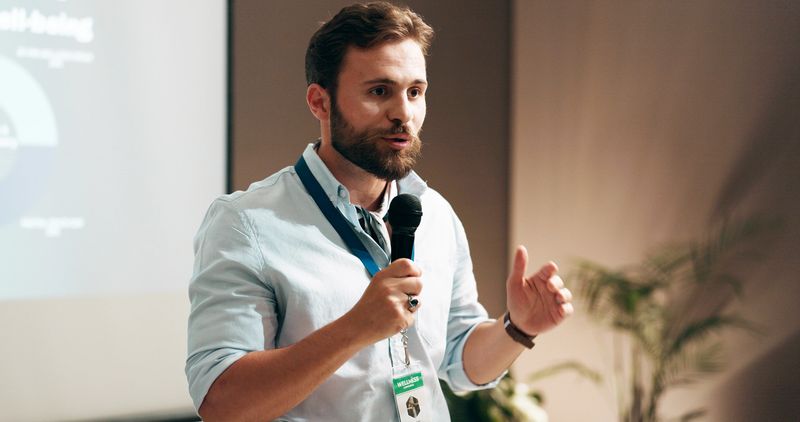 Businessman, development and presentation with speaker in conference room for wellness training. Education, forum speech and microphone with corporate coach on stage for public relations or seminar