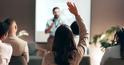 Person raising hand during a presentation in a conference room. billing accuracy, front desk operations, medical training, patient intake best practices, DX codes in PT, OT, and SLP