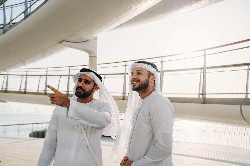 Two arab businessmen wearing traditional kandura are discussing and pointing while standing on a bridge in Dubai
