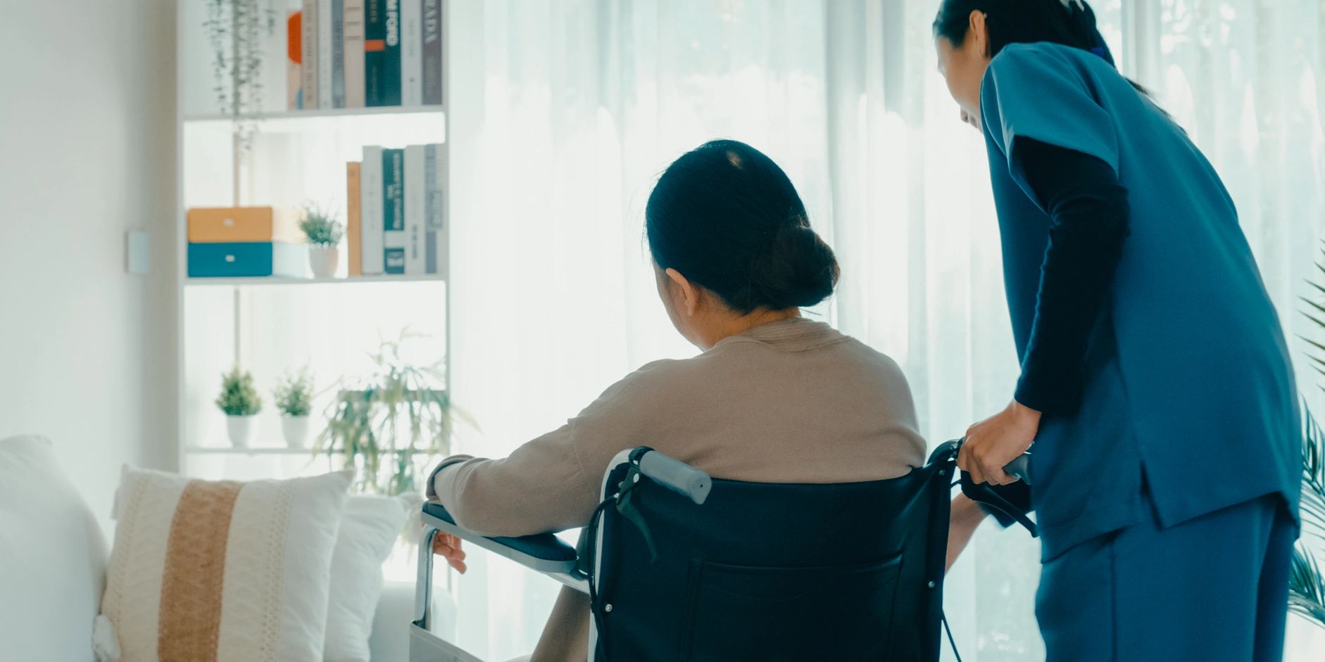 Caregiver assisting elderly woman in a wheelchair in a bright, cozy room.