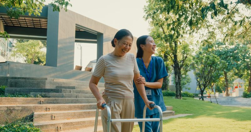 Young Asian nurse helps an elderly woman with a walker enjoy support and active living in a park. Outdoor health care concept.