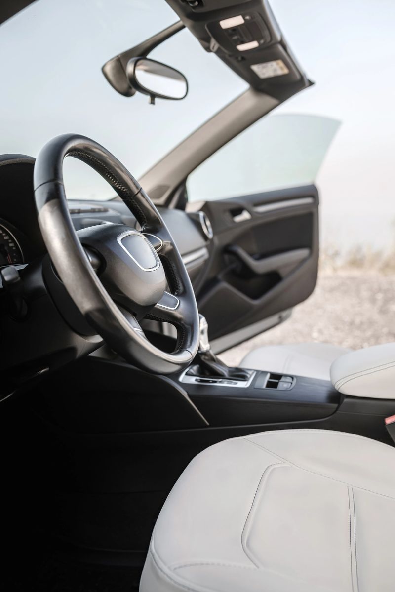 Luxury car interior with open door, white leather seats, steering wheel, and modern dashboard. Natural daylight, outdoor view, no people, vertical shot, sleek automotive design.