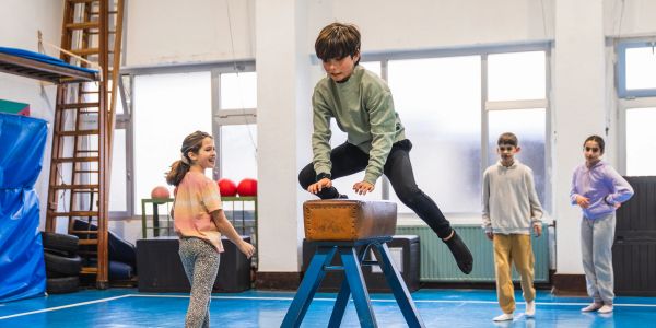 A boy jumps over gym equipment while others watch in a gymnasium.