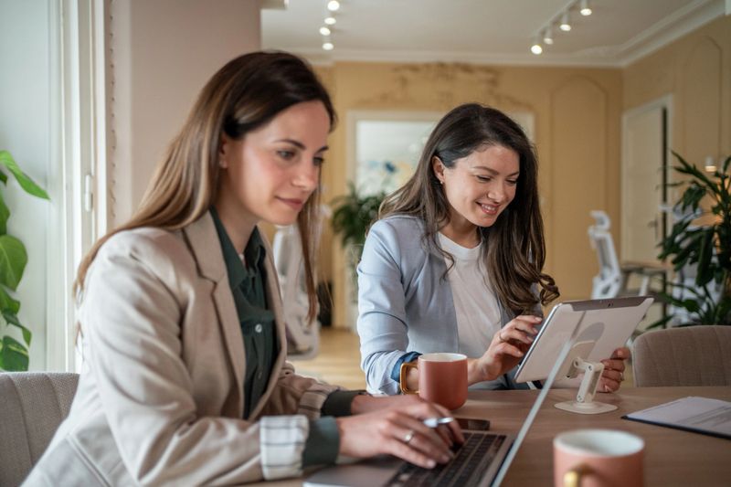 Two young businesswomen are working together on a project, using a laptop and a digital tablet in a modern office setting, sharing ideas and information