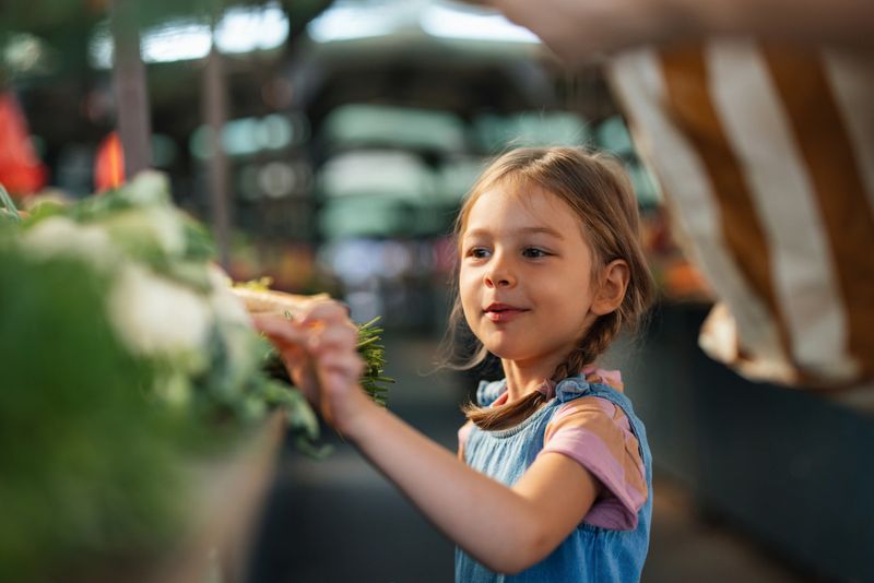 A cheerful young girl inspects fresh produce at a local outdoor market, enjoying a lively shopping experience surrounded by vibrant colors and aromas.
