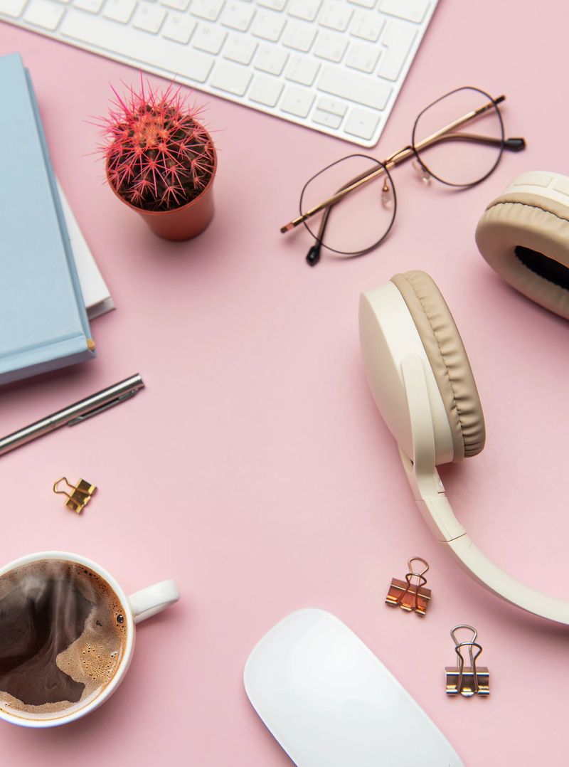 Home office desk workspace with keyboard, headphones, glasses, coffee, mouse, notebook and cactus plant on pink background