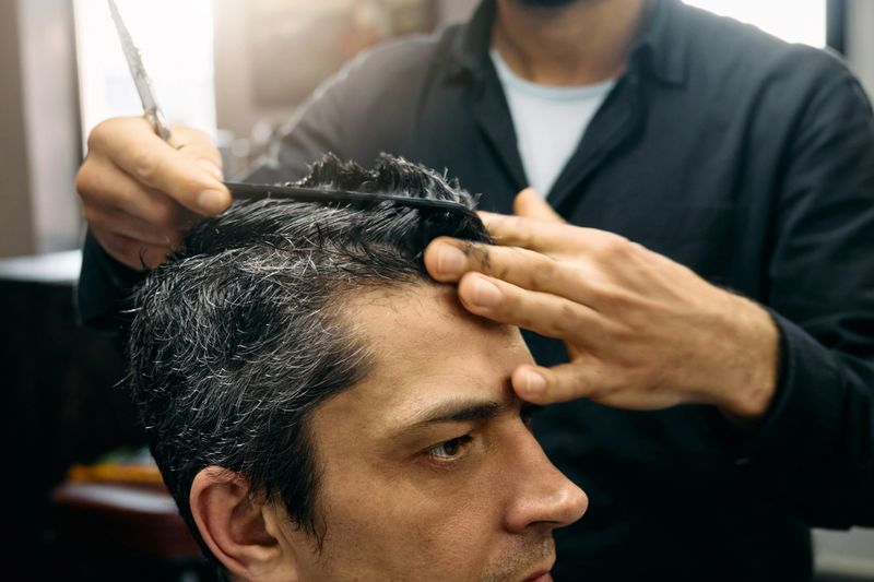 A skilled hairdresser is focused on giving a precise haircut to a client in a welcoming barbershop environment during a bustling afternoon. The stylist uses scissors and a comb for the trim.
