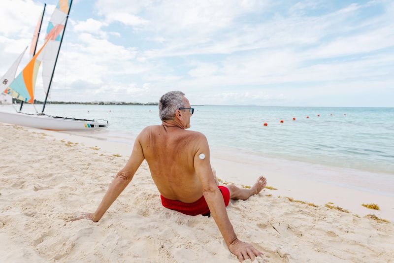 A senior man enjoys a serene moment on the beach, gazing at the ocean. The clear skies, calm waves, and bright catamaran sails enhance the atmosphere of relaxation and leisure.