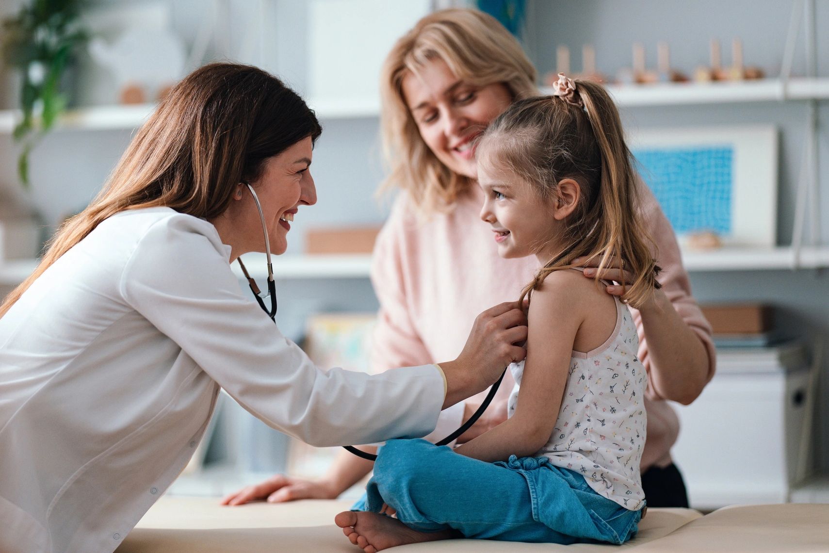 Doctor using a stethoscope to check a little girl's chest, with her mother smiling nearby.