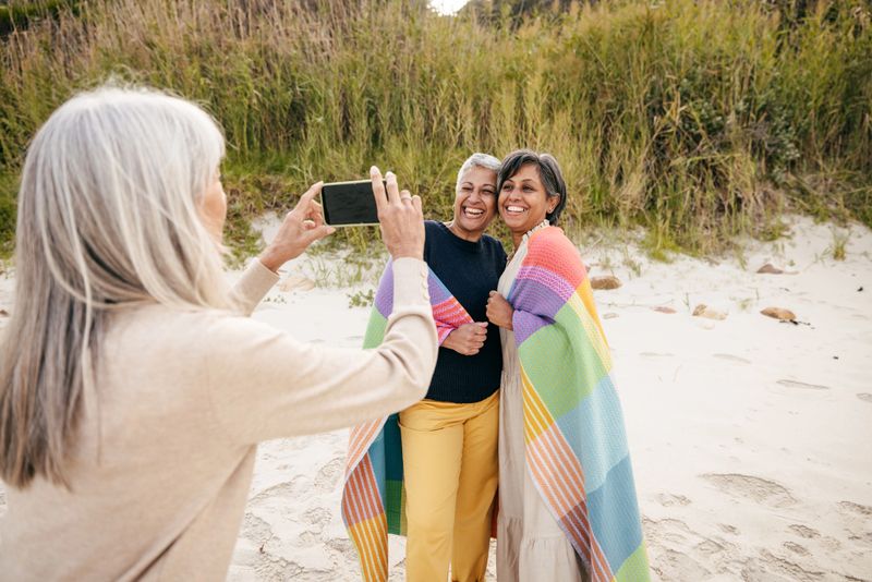 Two happy women enjoy a beachside moment, wrapped together in a vibrant striped blanket as their friend captures the memory.