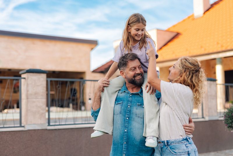 Smiling parents carrying their daughter on shoulders next to their new home