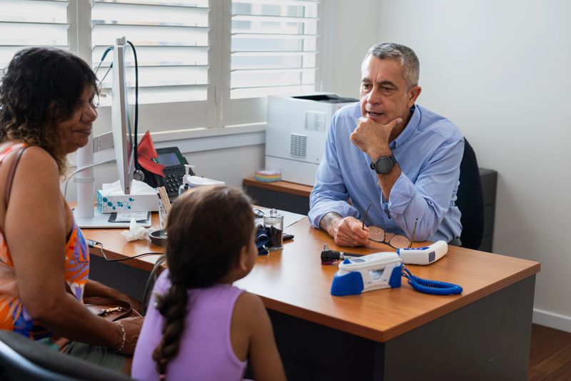 Middle aged Pacific Islander doctor consults with young Aboriginal patient and her Grandmother in his surgery