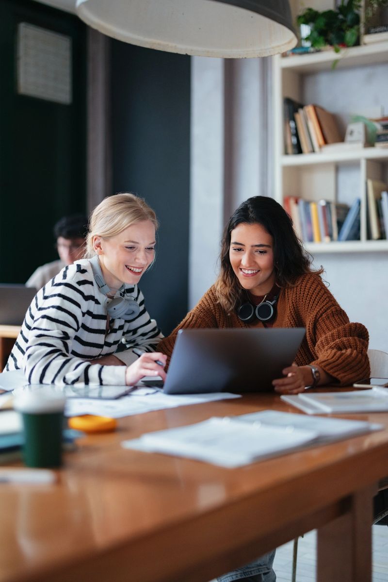 Two female students working on a laptop together, studying in a library environment with bookshelves and a modern decor in the background. The atmosphere highlights collaboration, learning, and academic pursuits.