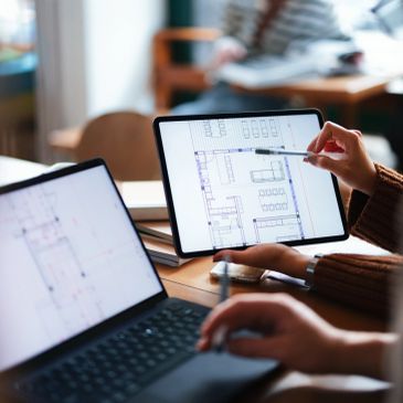 Person reviewing architectural plans on a tablet next to a laptop.