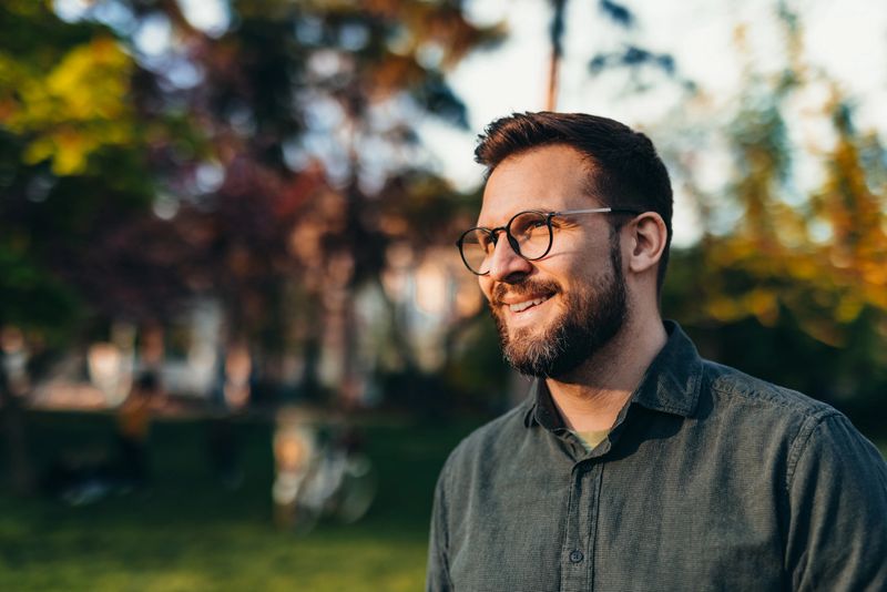 Smiling young man with eyeglasses looking away in a park during sunset, radiating confidence and happiness while surrounded by the warm glow of autumn trees and nature
