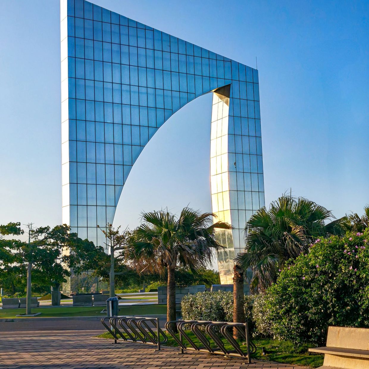 Modern glass arch structure with palm trees and clear blue sky.