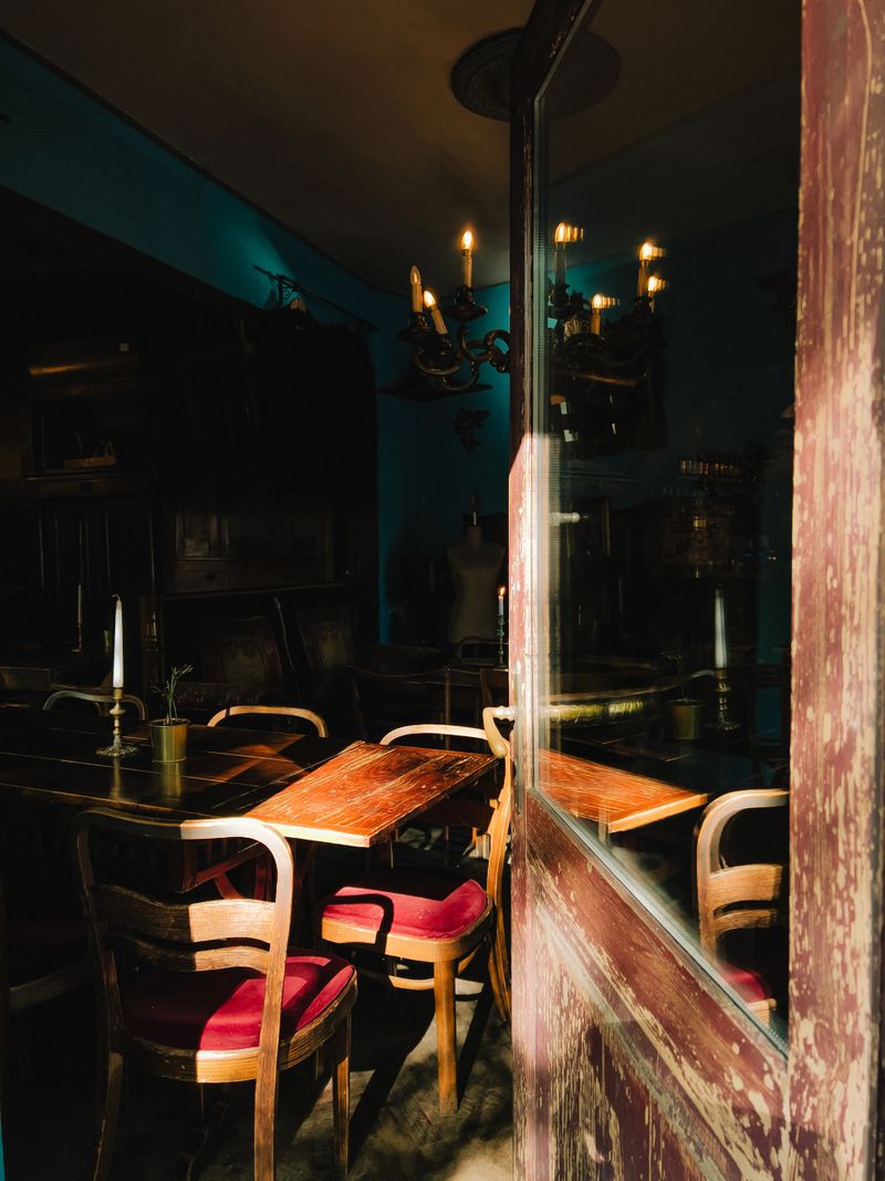 Sunlight streaming through weathered wooden door illuminates wooden tables and red upholstered chairs in cozy, vintage style restaurant, creating warm ambiance. Red chairs inside vintage restaurant