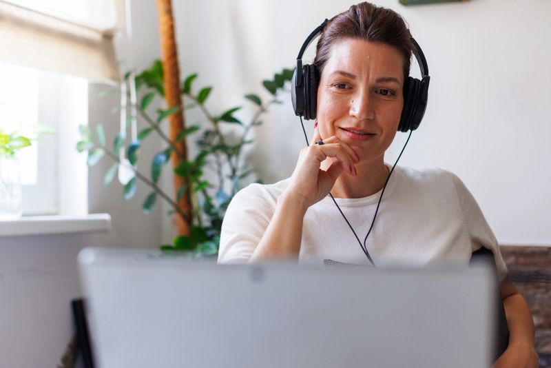 Focused woman wearing headphones sits at a table, attending a virtual meeting or video conference on her laptop. She gestures with her hands while communicating, suggesting active participation. The home interior features modern decor, natural lighting, and green plants, illustrating a productive remote work environment.