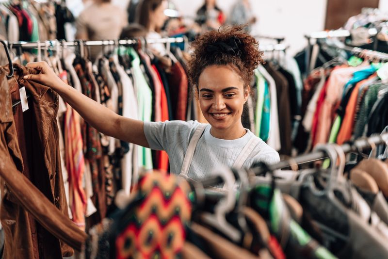Young woman smiling while browsing through clothes in a second-hand shop, embracing sustainable fashion and promoting a zero-waste lifestyle with each thoughtful choice