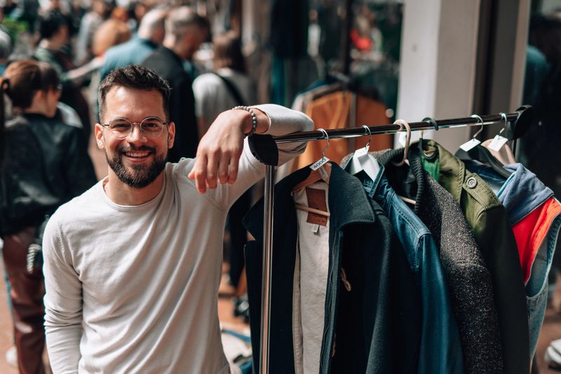 Young adult male vendor smiling at the camera while leaning casually against a clothing rack in an outdoor market, showcasing stylish second-hand fashion and creating a friendly atmosphere