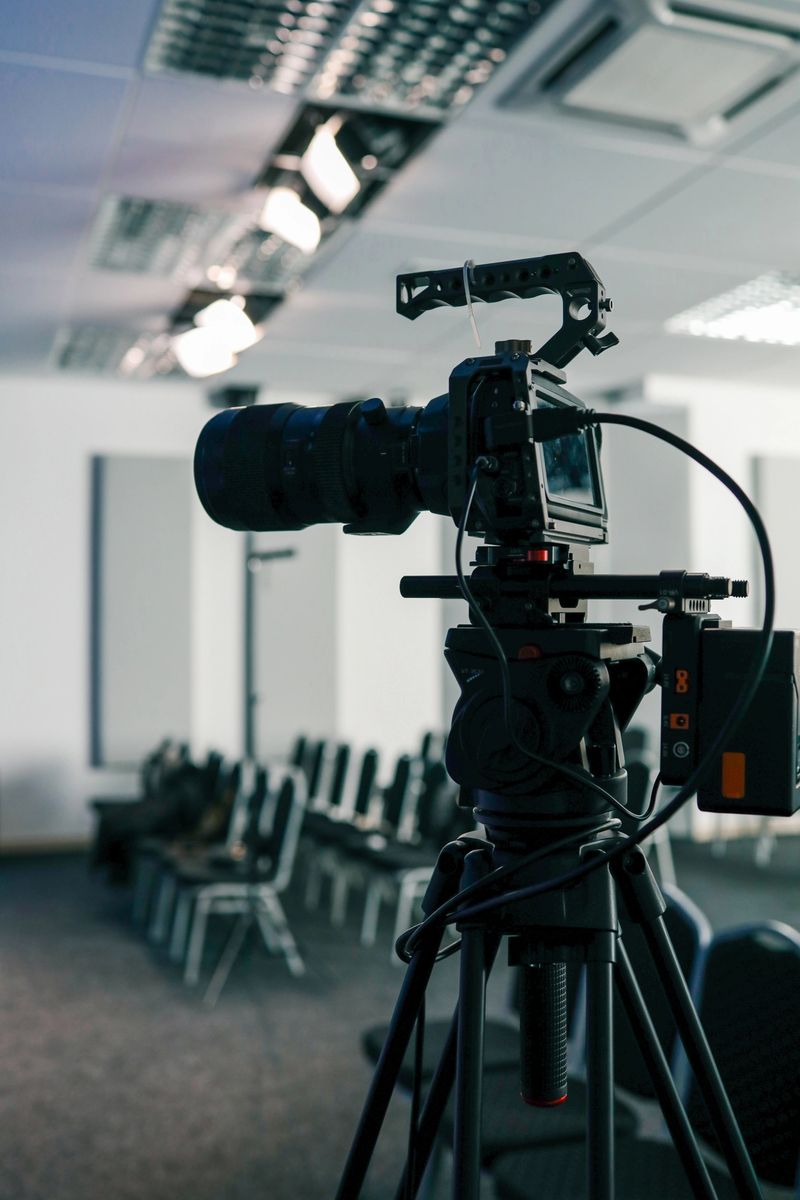 Professional video camera on a tripod set up in a modern conference room, ready for filming or live streaming, with rows of empty chairs and ceiling lights in the background.