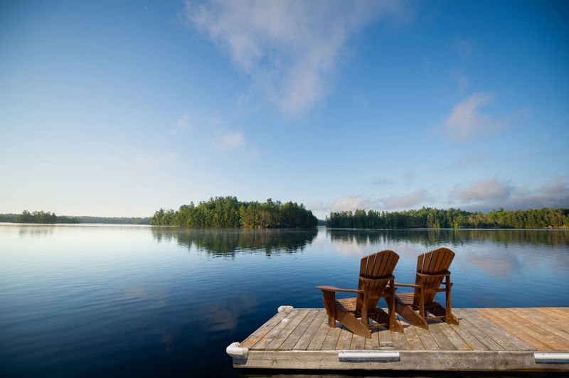 Two Adirondack chairs sit on a wooden dock overlooking a calm Muskoka lake, with fluffy clouds and lush green trees mirrored in the still water. Space for copy.