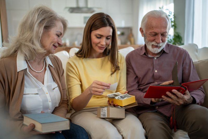 Smiling parents and their adult daughter reminiscing while sharing old memories from a photo album, sitting comfortably on the sofa in the cozy living room at home