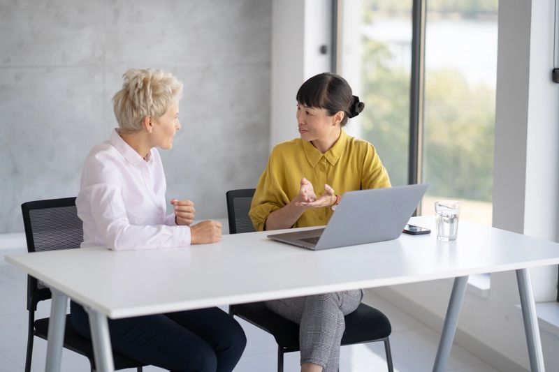 A productive exchange of ideas occurs as two women share insights using a laptop in a bright, open office environment.