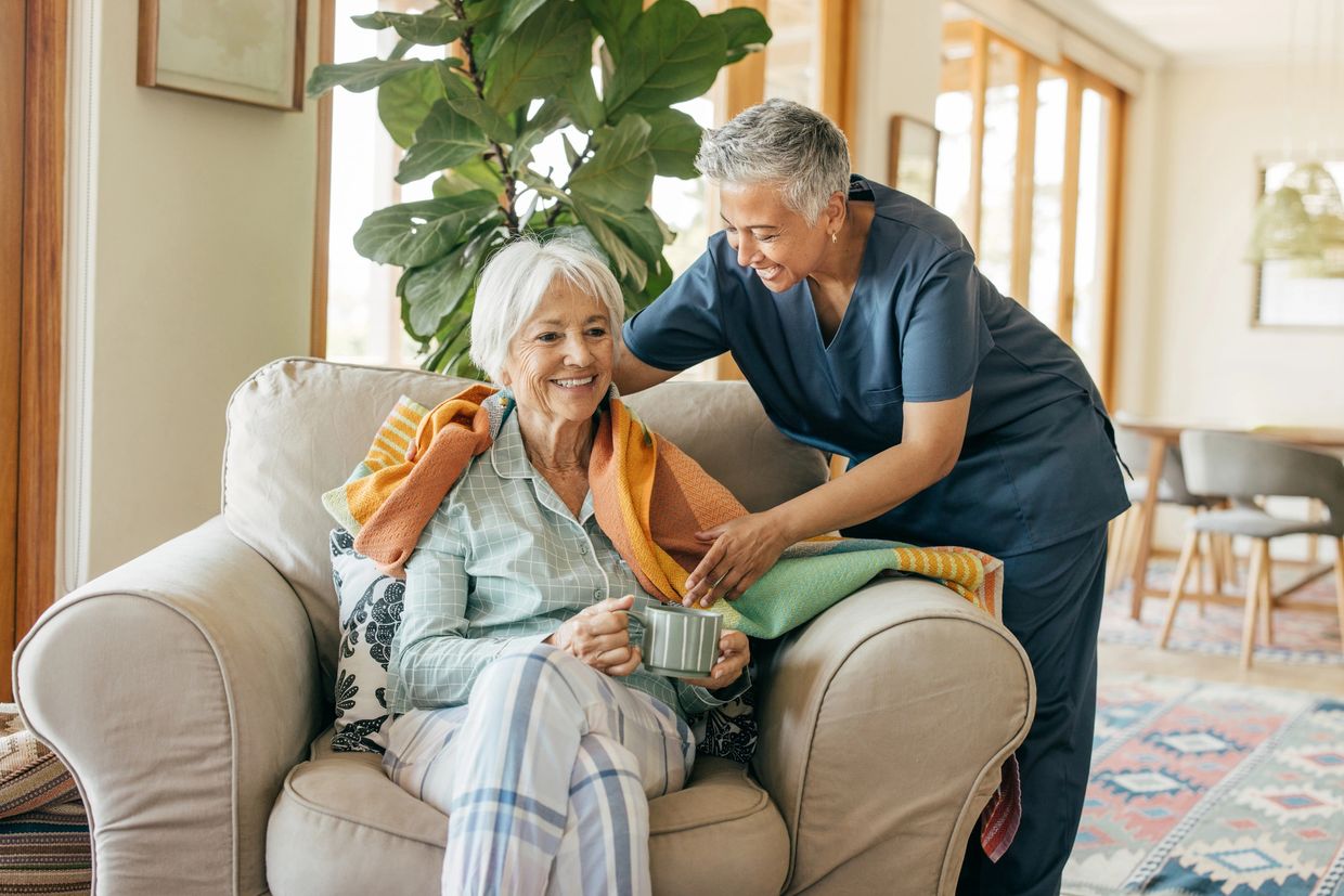 Caregiver wrapping a warm blanket around a smiling elderly woman on a cozy armchair.