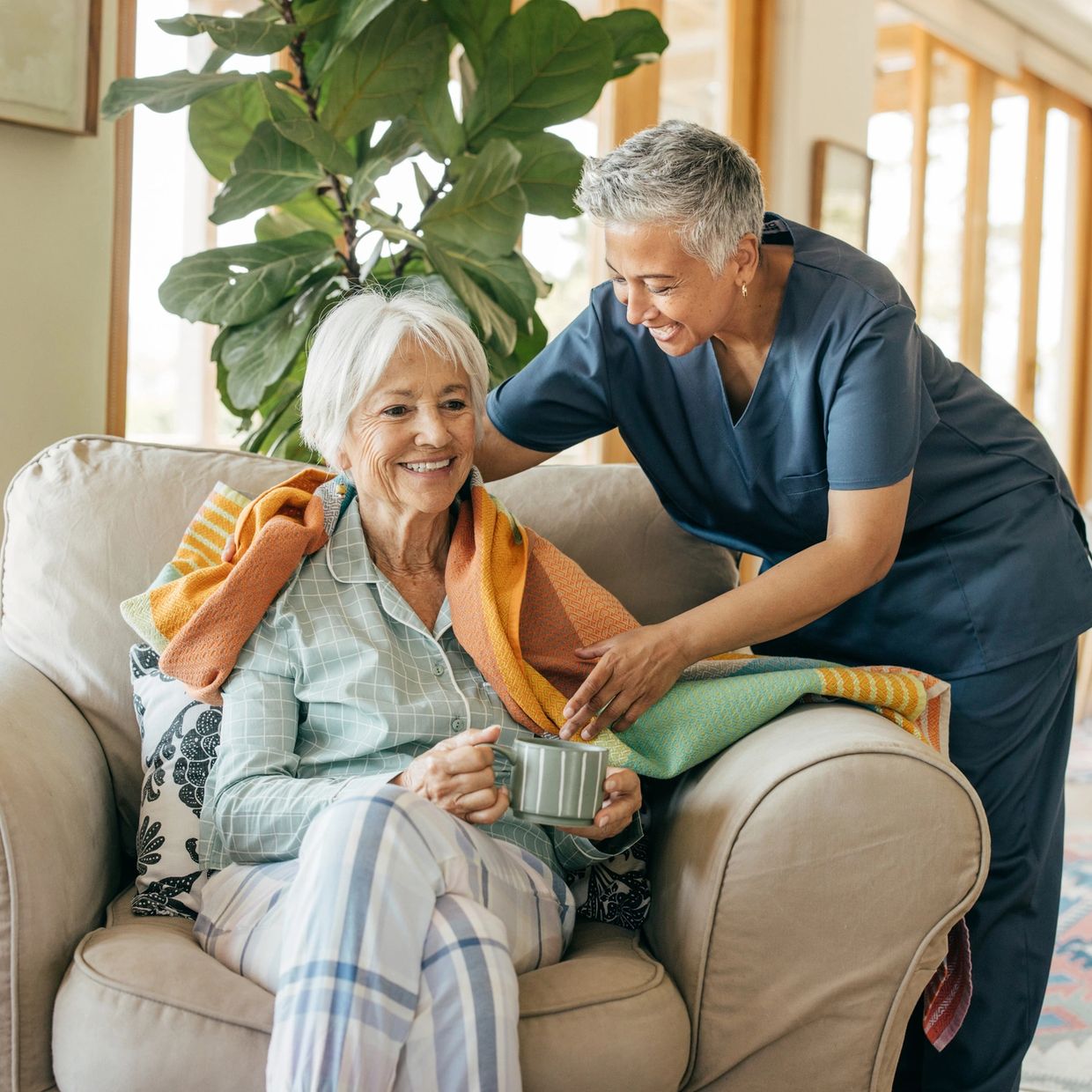 Caregiver warmly covering elderly woman with a blanket as she holds a cup.