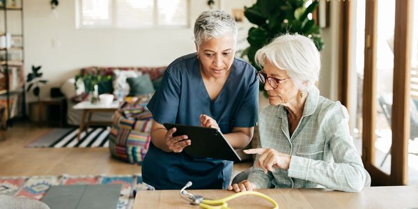 A healthcare worker assists an elderly woman with a tablet in a cozy home setting.