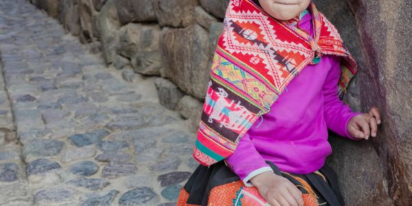 A young child in colorful traditional clothing sitting on a stone path beside a stone wall.