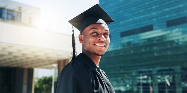 Smiling graduate in black cap and gown outdoors on a sunny day.