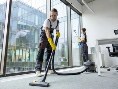 Two janitors cleaning an office with a vacuum and window cleaner.