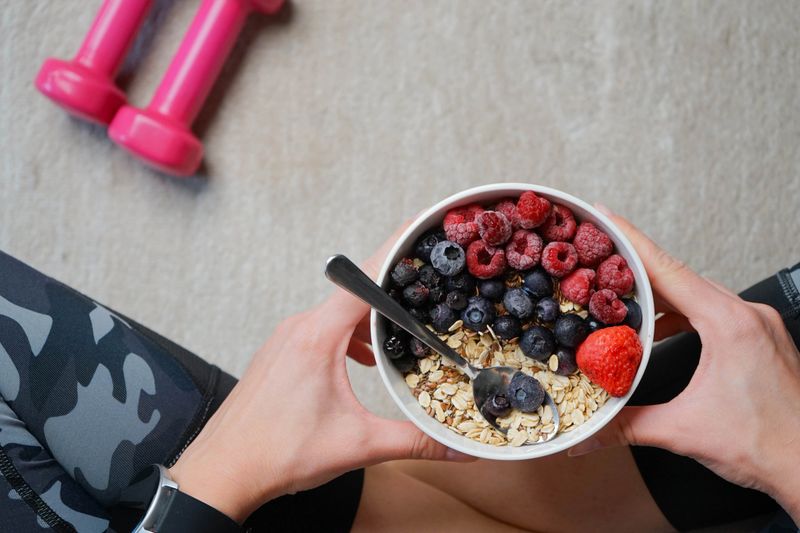 After her fitness training, a girl sits on her exercise mat at home, eating a bowl of oats with forest fruits. Holding the bowl in her hand, she enjoys the healthy mix of oats and fresh berries—blueberries, raspberries, and strawberries. Her face shows a sense of fulfillment and relaxation. The warm and inviting atmosphere of the room makes it the perfect space for a rejuvenating post-workout snack.