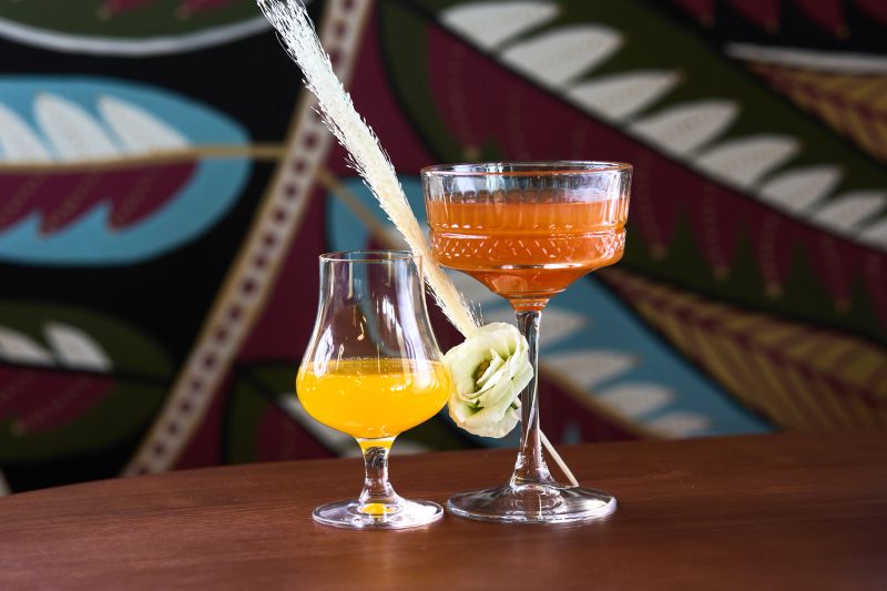 A visually stunning pair of cocktails, featuring a vibrant orange cocktail in a textured coupe glass and a bright yellow cocktail in a curved glass. The orange cocktail is garnished with a delicate white flower, while the yellow cocktail has a decorative feather. Set against a colorful background with tropical patterns, these beverages exude elegance and sophistication, perfect for a luxurious event.