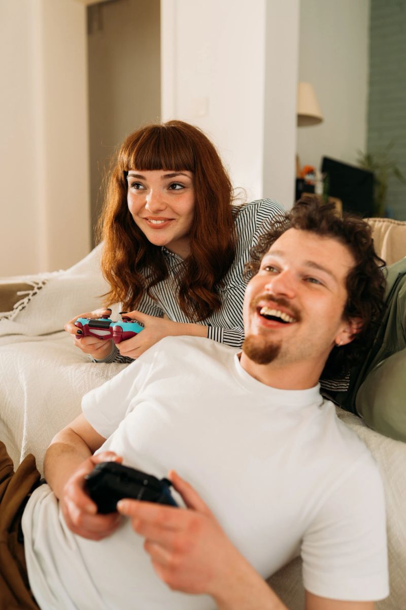 A young couple sits close on the sofa, playing video games and having a blast. He’s smiling as he concentrates on the game, and she’s laughing, clearly having fun. Their controllers match their energy ,bright and playful.