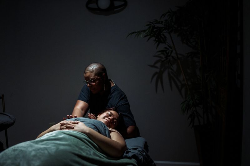 A beautiful Hispanic woman lays on her back on a massage table in a dimly lit treatment room at a luxury spa and smiles with pleasure while a Black female massage therapist massages on her neck and shoulders.
