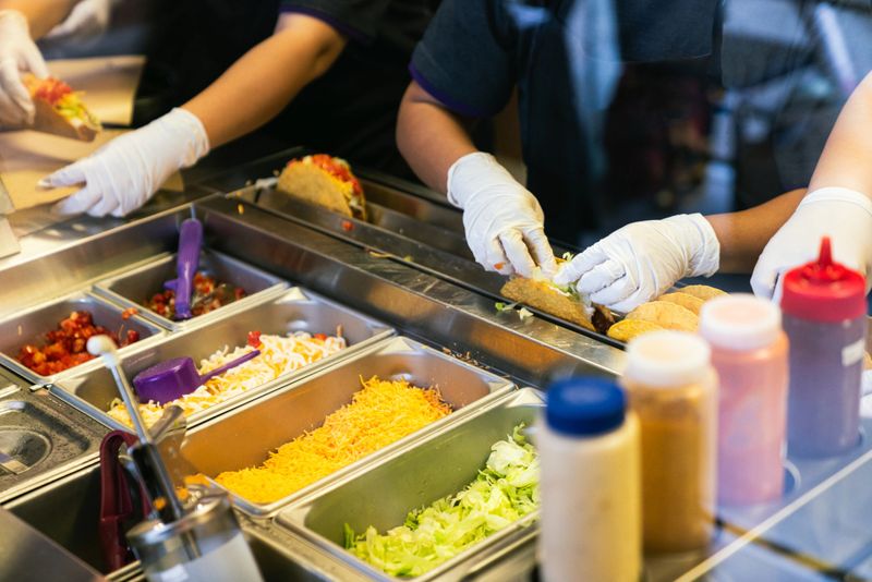 A close-up of food service workers preparing Mexican tacos at a fast food restaurant, with gloved hands assembling tacos using various fresh ingredients on a stainless steel counter.